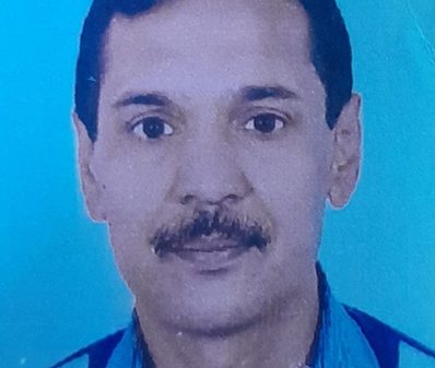 Portrait of a man with short dark hair and mustache wearing a blue shirt against a blue background.