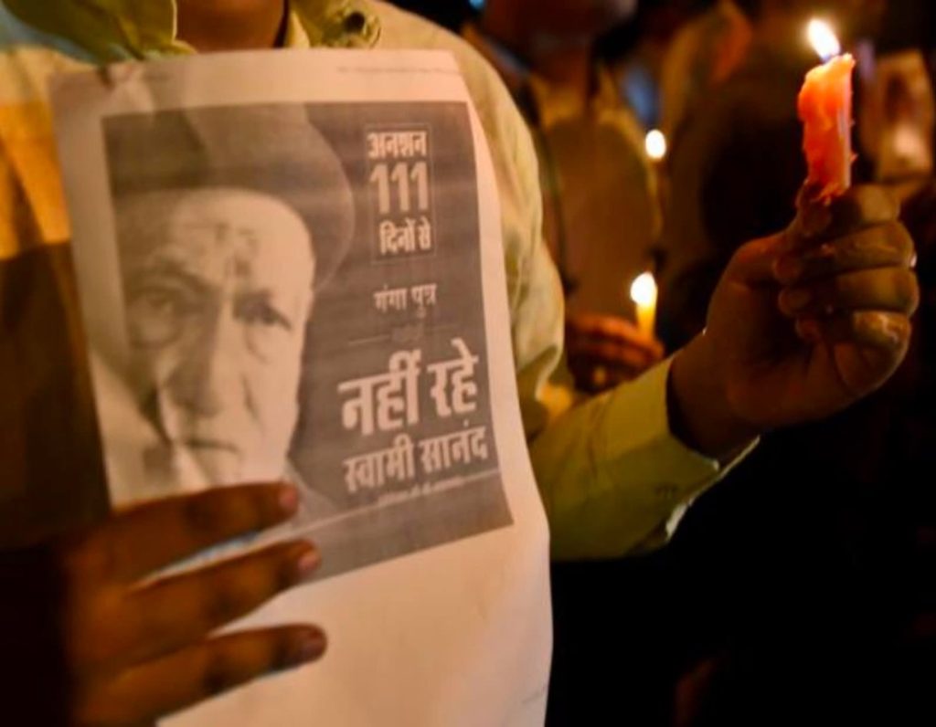 swami sanand | No. 1 Indian Media News Portal Person at night holding a poster with Gandhi's image while another hand holds a lit candle.