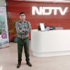 Man standing with arms crossed in a NDTV reception area, red wall with a large NDTV logo and a white curved desk behind him.