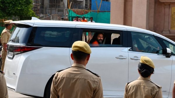 Bearded man in a white van waves from the window while security personnel in khaki uniforms stand nearby.