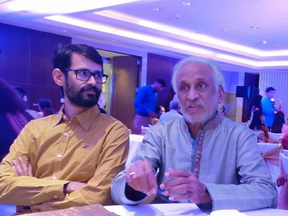 vijay sanghvi | No. 1 Indian Media News Portal Two men sit at a banquet table under blue lighting; an older man in a light gray kurta gestures while speaking to a younger man in a mustard shirt and glasses.