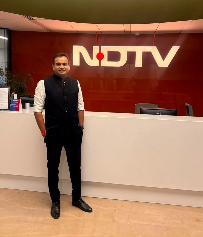 vivek shukla | No. 1 Indian Media News Portal Man in a black vest and white shirt stands at a white reception desk in a NDTV office lobby, with a lit NDTV logo on a red wall behind.