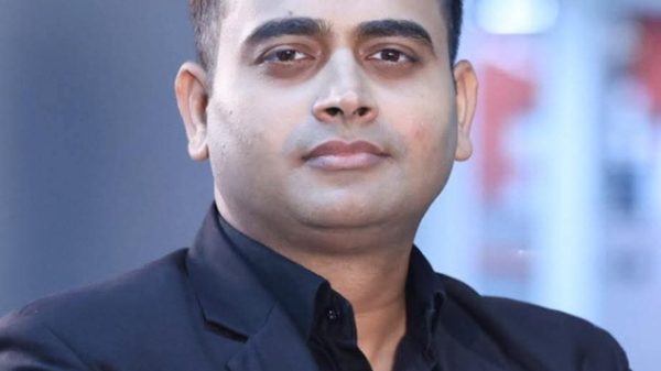 Professional headshot of a man in a black suit looking at the camera against a blurred background.