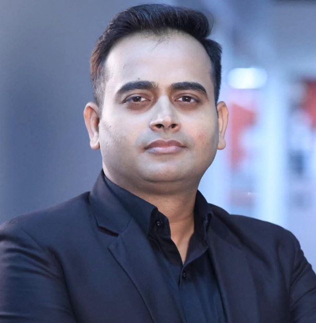 Professional headshot of a man in a black suit looking at the camera against a blurred background.