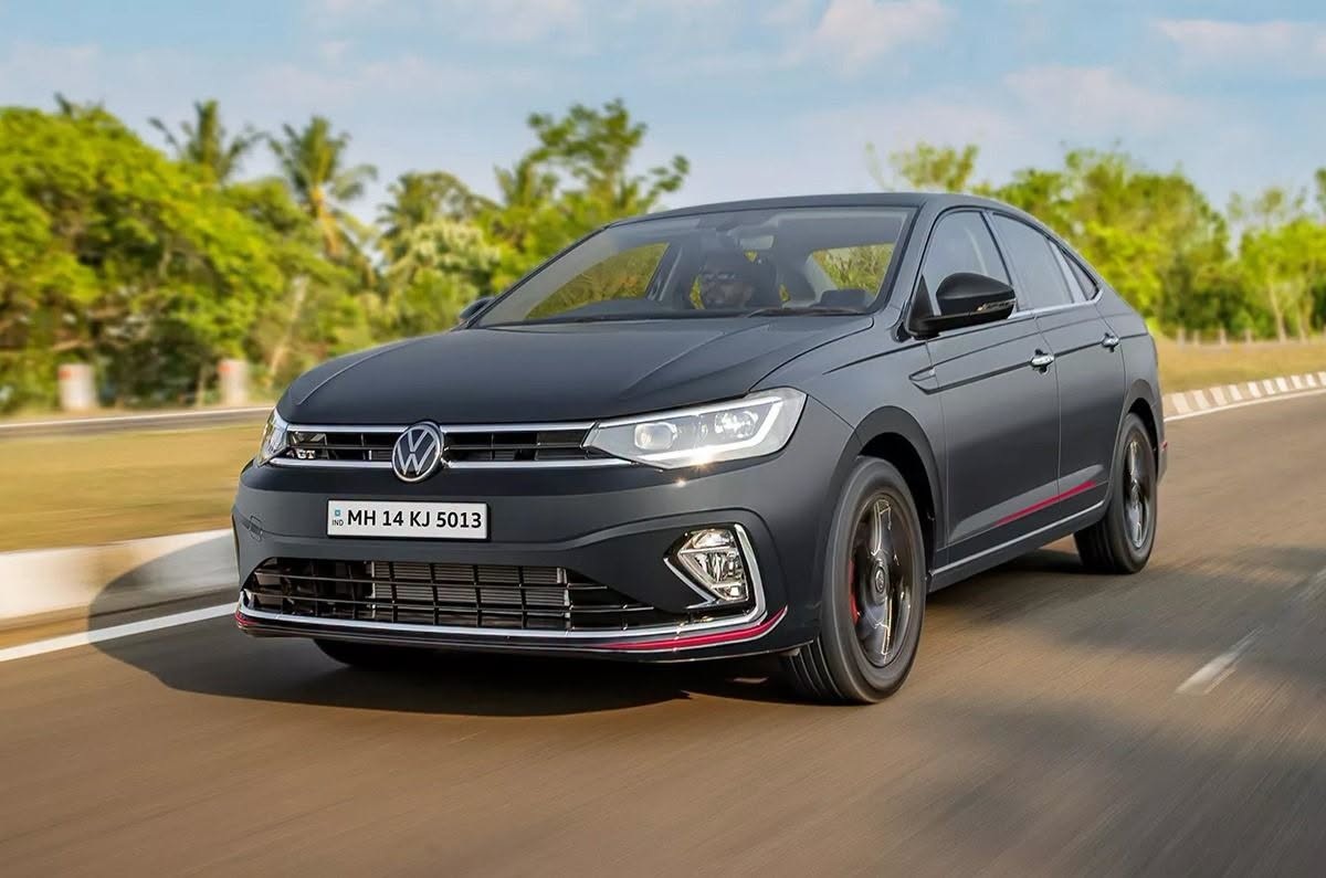 Gray Volkswagen sedan driving on a sunlit road with trees in the background (front three-quarter view)./ Rearview mirrors visible, motion blur suggests speed.