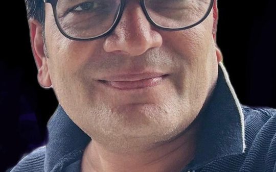 Close-up of a smiling man wearing black-rimmed glasses and a navy zip-up sweater against a dark background.