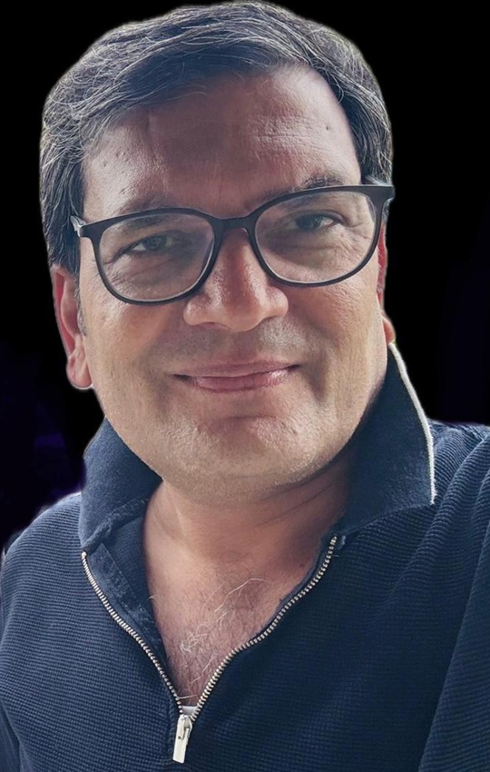 Close-up of a smiling man wearing black-rimmed glasses and a navy zip-up sweater against a dark background.