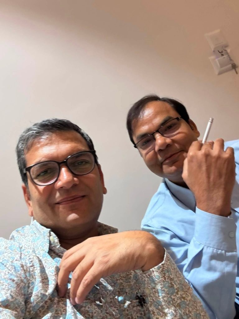 Two men wearing glasses take a selfie indoors; one in a patterned shirt, the other in a light blue shirt holding a pen.