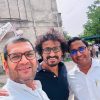 Three men wearing glasses posing for a selfie outdoors, with a concrete building and trees in the background.