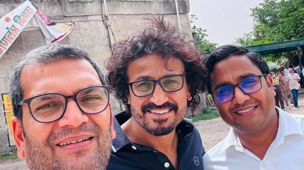 Three men wearing glasses posing for a selfie outdoors, with a concrete building and trees in the background.