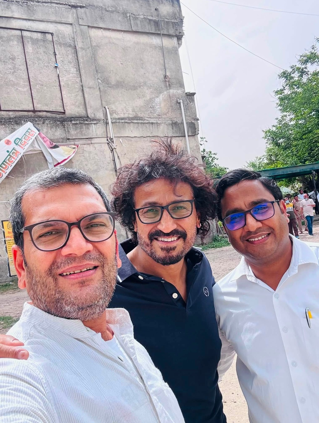 Three men wearing glasses posing for a selfie outdoors, with a concrete building and trees in the background.