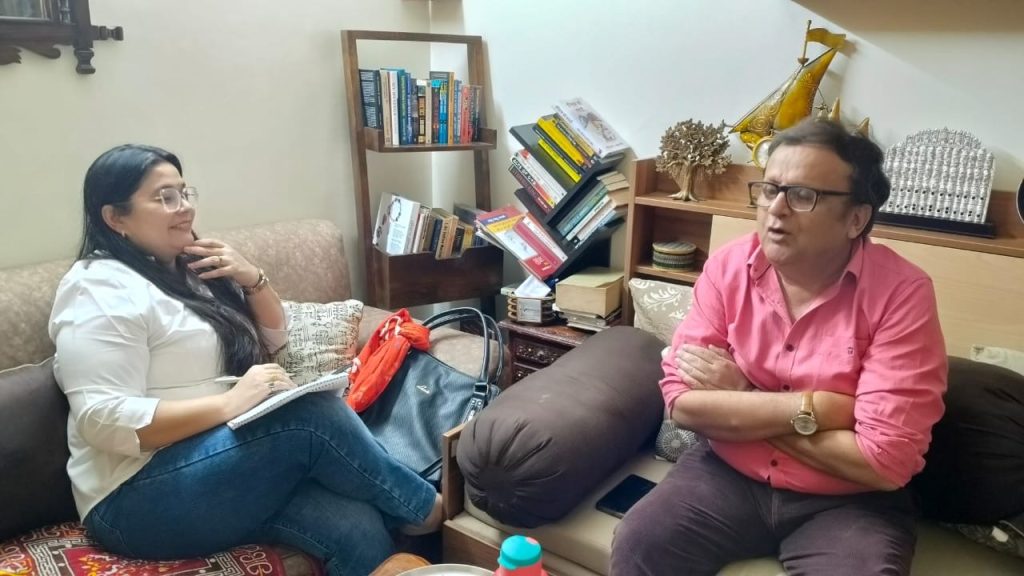 Woman in a white blouse sits on a couch with a notebook, talking to a man in a pink shirt.
