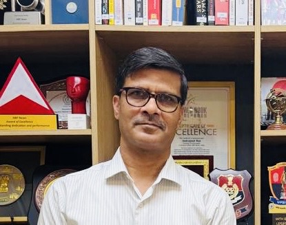 Man wearing glasses and a white shirt stands in front of shelves filled with awards and trophies.