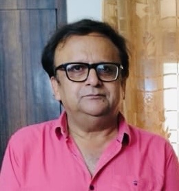 Man with dark hair and glasses wearing a pink shirt, standing indoors beside a blue door and sheer curtain.