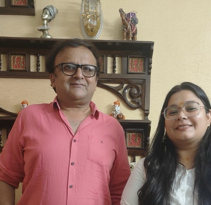 Man in pink shirt and glasses and a woman with long hair and glasses stand indoors in front of a carved wooden shelf with decorative figurines.