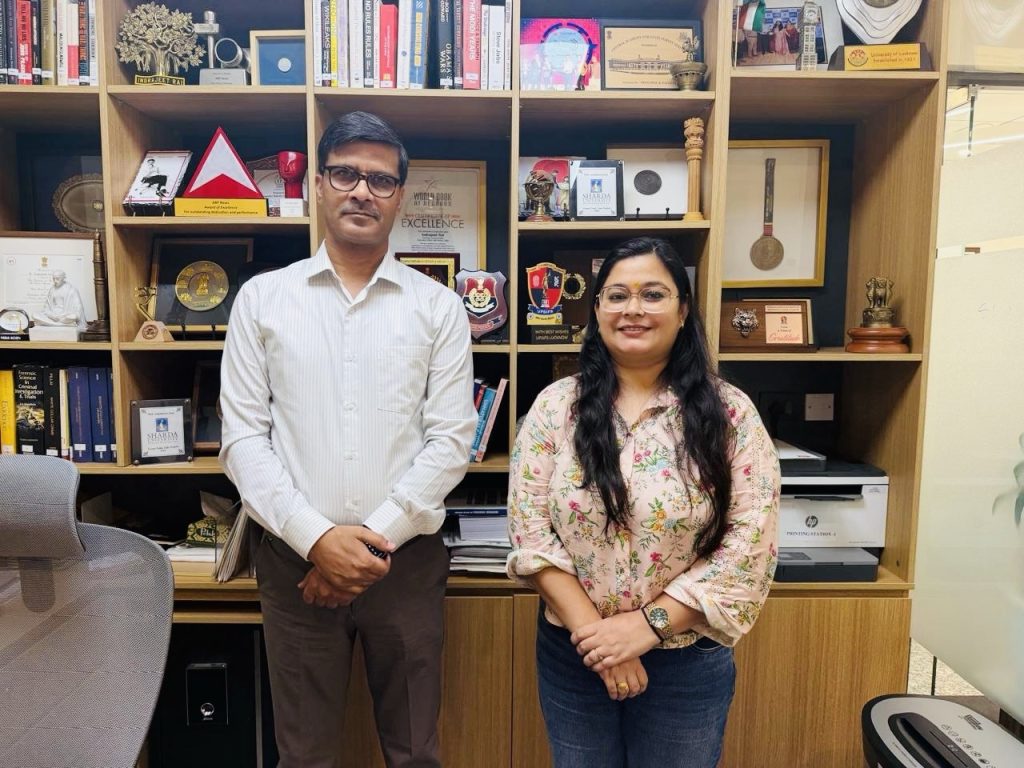 52ddc60f-7ab9-4afa-967b-8e0ffa0c2259 | No. 1 Indian Media News Portal Two professionals stand side by side in an office with wooden shelves filled with awards and books behind them.