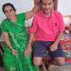 An elderly woman in a green sari blessing a young man with a red tika on his forehead, indoors on a colorful cloth-covered bench table.