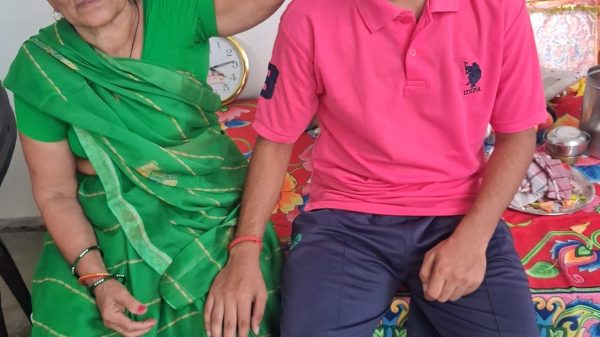 An elderly woman in a green sari blessing a young man with a red tika on his forehead, indoors on a colorful cloth-covered bench table.
