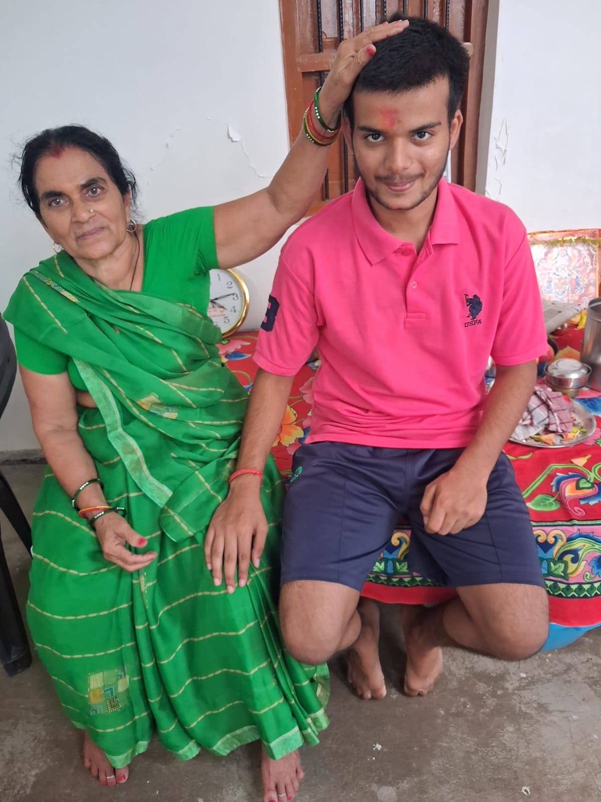 An elderly woman in a green sari blessing a young man with a red tika on his forehead, indoors on a colorful cloth-covered bench table.