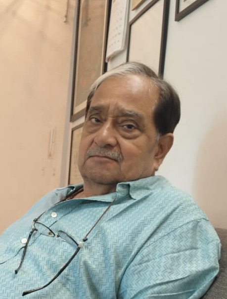 628f2244-65d2-4d82-b422-4b4247ba4930 | No. 1 Indian Media News Portal Older man seated indoors, wearing a turquoise patterned shirt and glasses hanging from his chest, looking at the camera.