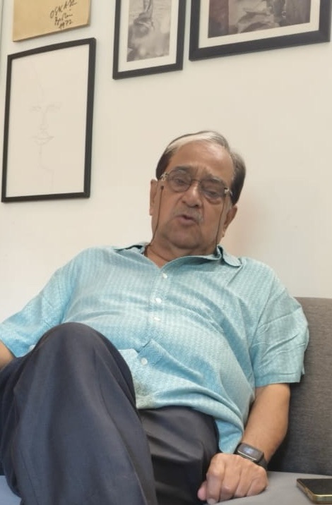 7c82a4c9-28d3-4429-ad8b-84c4abe7b134 | No. 1 Indian Media News Portal Older man with glasses in a light blue shirt sitting on a gray sofa, leaning back, with framed photos on the wall behind him.