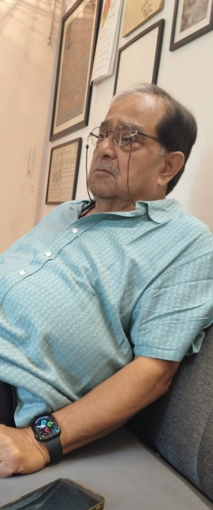 9ab5b66d-33c1-484b-b673-a0224bdd5528 | No. 1 Indian Media News Portal Older man wearing a turquoise patterned shirt sits on a sofa, looking down, wearing glasses and a smartwatch.