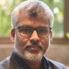 Portrait of a middle-aged man with gray hair and beard wearing glasses, looking at the camera with a blurred indoor background and a plant.