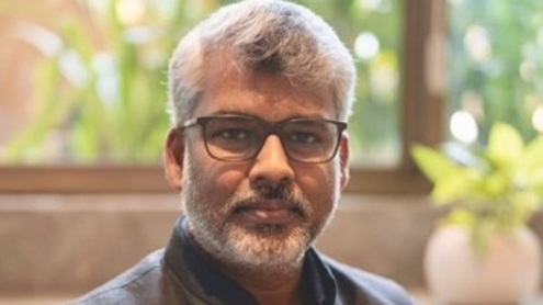 Portrait of a middle-aged man with gray hair and beard wearing glasses, looking at the camera with a blurred indoor background and a plant.