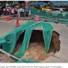 Large sinkhole in a city street, tarps and orange traffic cones cordon off the hole while onlookers stand behind a barrier.