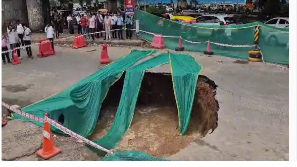 Large sinkhole in a city street, tarps and orange traffic cones cordon off the hole while onlookers stand behind a barrier.