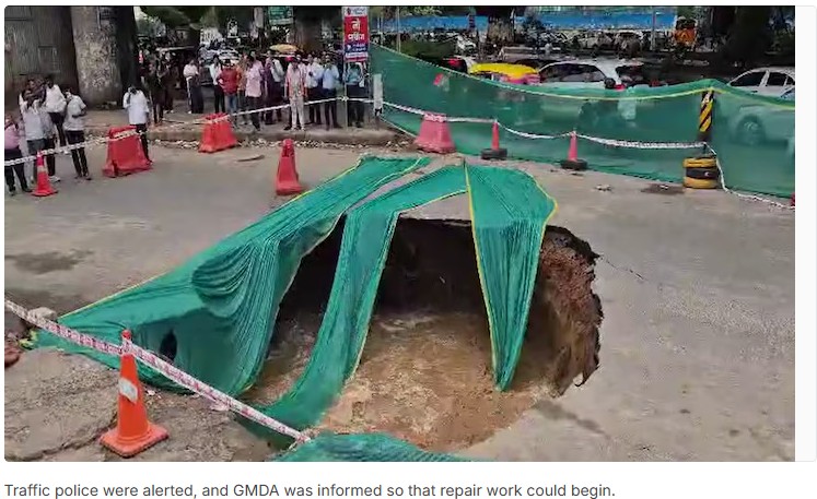 Large sinkhole in a city street, tarps and orange traffic cones cordon off the hole while onlookers stand behind a barrier.
