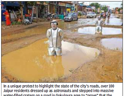 Astronaut-costumed protester wades through muddy floodwater on a Jaipur street, highlighting road craters in a local protest.