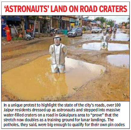 Astronaut-costumed protester wades through muddy floodwater on a Jaipur street, highlighting road craters in a local protest.
