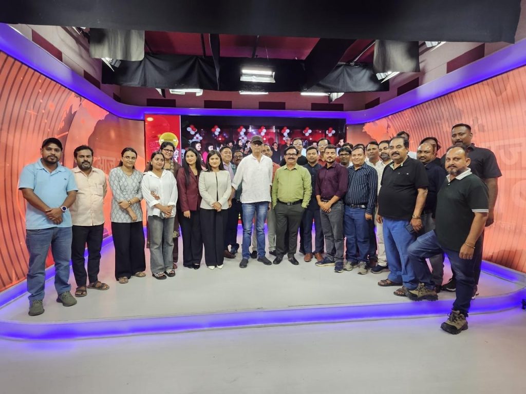 Large group of people standing together on a TV studio set with orange walls and purple lighting.
