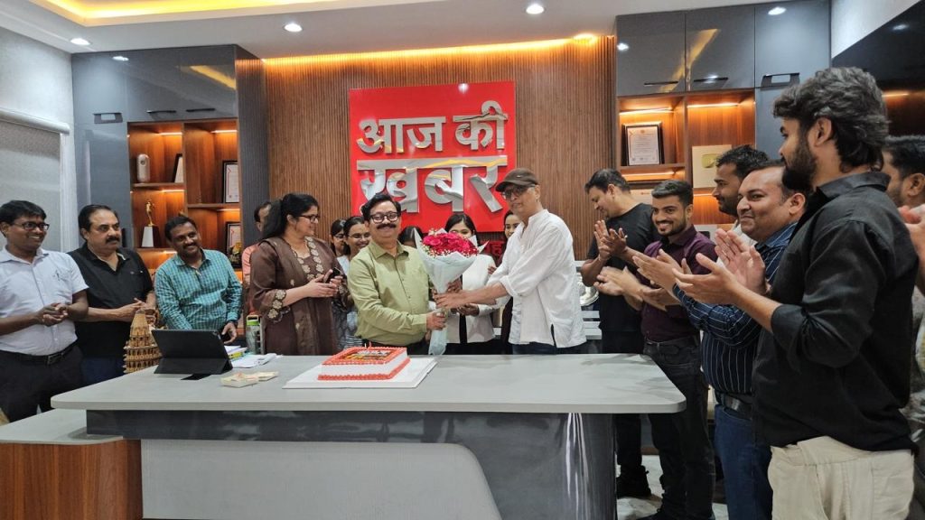 Group of coworkers at a celebration around a modern office desk; a man hands a bouquet to another while colleagues applaud, with a red 'आज की खबर' sign in the background and a cake on the table.