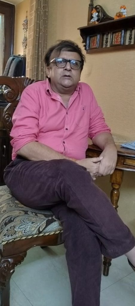 Man in a pink shirt wearing glasses sits on a wooden chair in a living room with a shelf and decorations behind him, looking toward the camera.