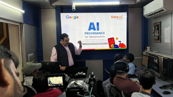 Presenter standing beside a large screen that reads 'AI Provenance for Newsrooms' with Google and Data LEADS logos; audience watches in a studio control room.