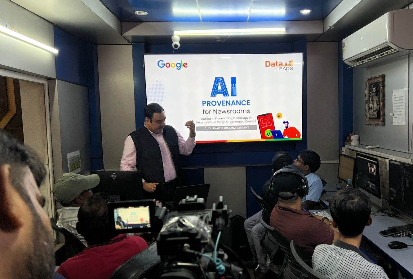 Presenter standing beside a large screen that reads 'AI Provenance for Newsrooms' with Google and Data LEADS logos; audience watches in a studio control room.