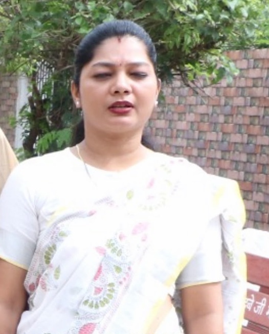akanksha yadav | No. 1 Indian Media News Portal Woman wearing a white saree with floral patterns, standing outdoors with green foliage and a brick wall behind her.