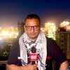 Male reporter with glasses and a black-and-white scarf holds a red news mic on a rooftop at night, city lights behind him.