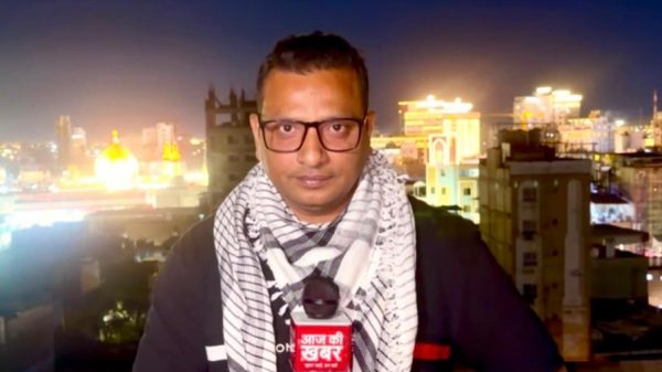 Male reporter with glasses and a black-and-white scarf holds a red news mic on a rooftop at night, city lights behind him.