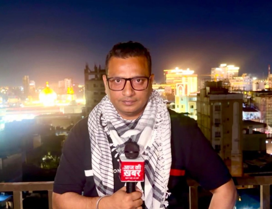 Male reporter with glasses and a black-and-white scarf holds a red news mic on a rooftop at night, city lights behind him.