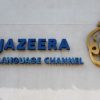 Al Jazeera English Language Channel sign with blue letters and a gold emblem on a light wall.