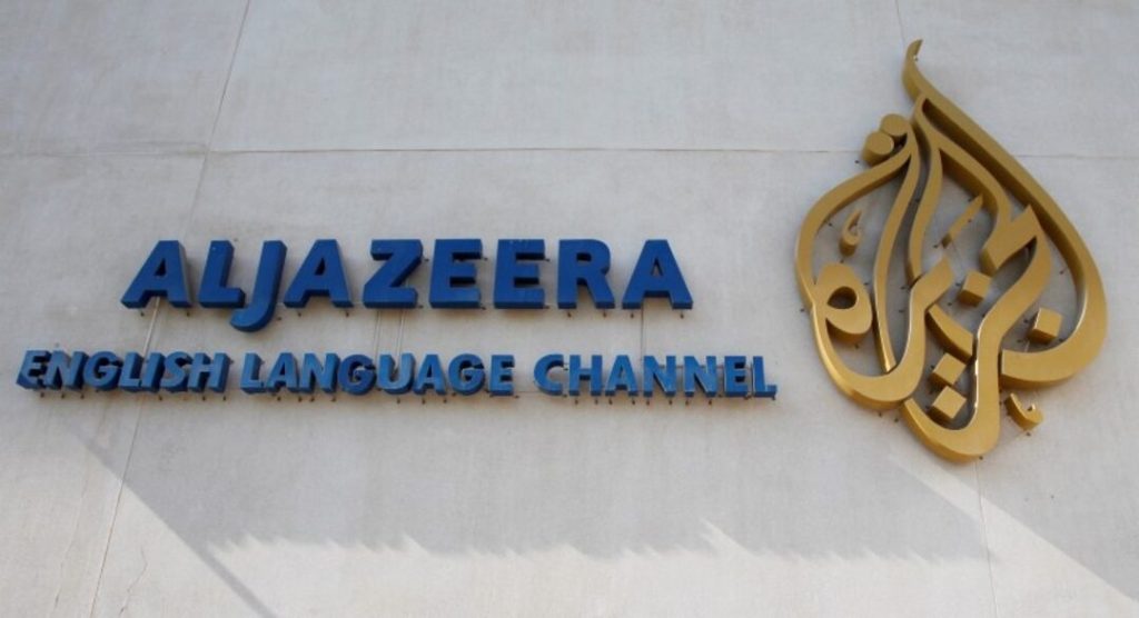 Al Jazeera English Language Channel sign with blue letters and a gold emblem on a light wall.