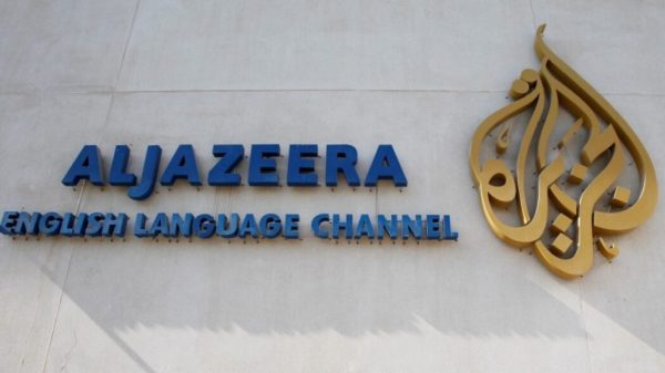 Al Jazeera English Language Channel sign with blue letters and a gold emblem on a light wall.