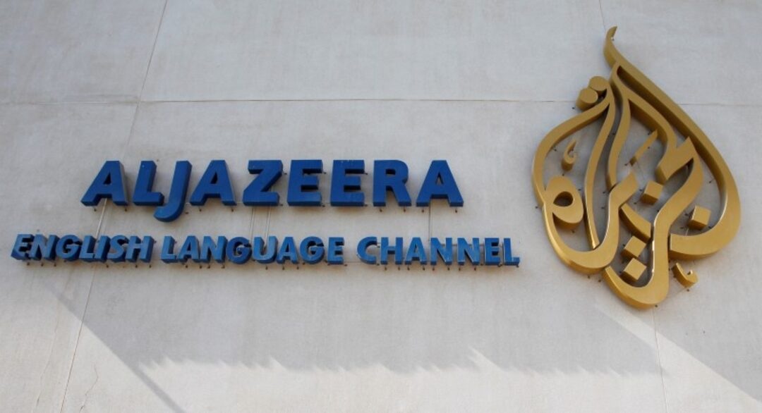 Al Jazeera English Language Channel sign with blue letters and a gold emblem on a light wall.