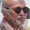 Older man with a gray beard and bald head, wearing round sunglasses and a brown plaid shirt, outdoors (backpack strap over his shoulder).