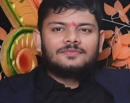 Portrait of a man with a red tilak on his forehead, wearing a dark blazer, smiling softly, against a colorful festive backdrop