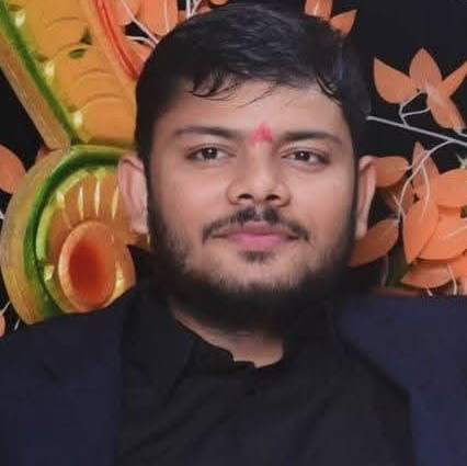 Portrait of a man with a red tilak on his forehead, wearing a dark blazer, smiling softly, against a colorful festive backdrop