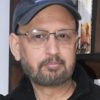 Portrait of a man wearing a dark cap and glasses, with a short beard, standing in front of a bookshelf.